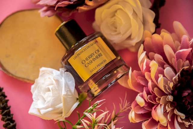 Une photo illustrative du parfum de niche Cherry of Cashmere entouré de 2 fleurs blanches et d'une fleur rose.