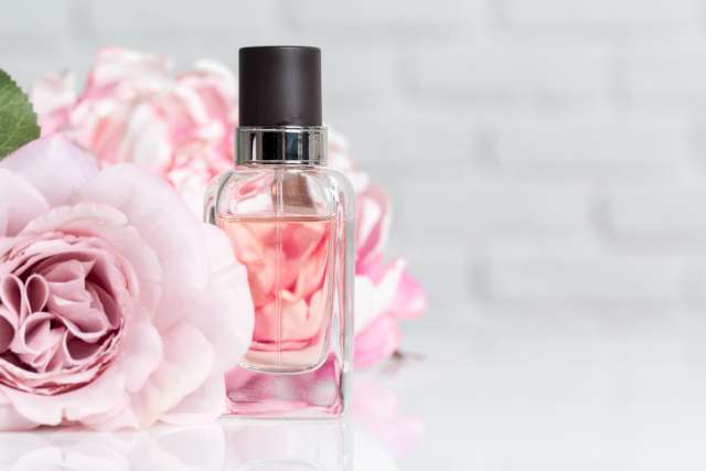 Une photo illustrative d'un flacon de parfum transparent mis a coté d'une fleur de rose. Le fond est flouté, à moitié gris et l'autre moitié est constituée par une deuxième toujours floutée.