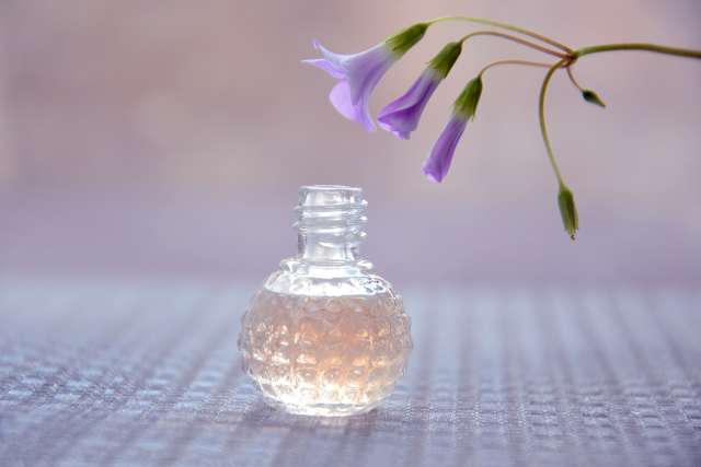 Une photo illustrative d'un flacon de parfum ayant une forme ronde et trois fleurs violette en dessus du flacon. 