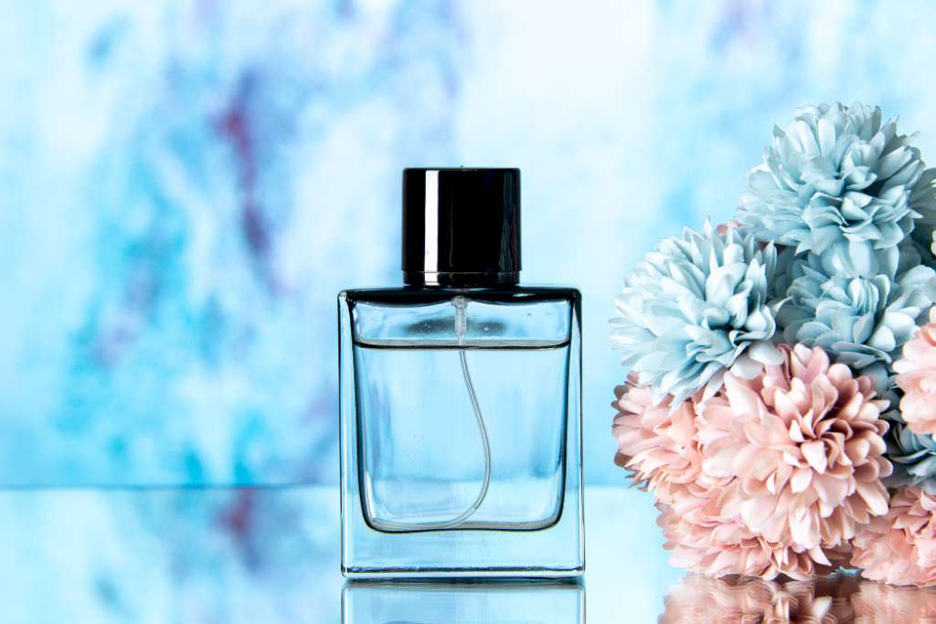 Une photo d'un flacon de parfum simple mis à coté de fleurs roses et bleus sur un fond bleu floutée.